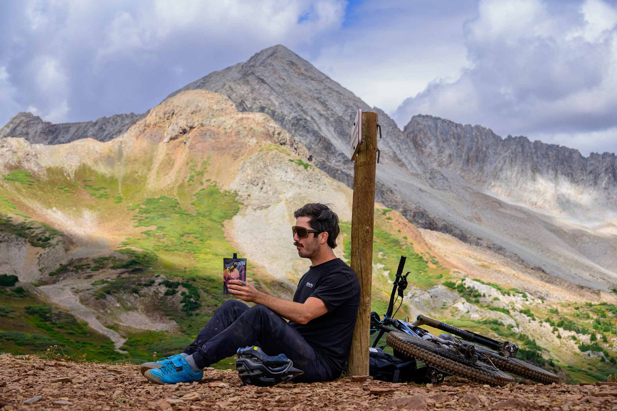 Shawn Neer sits against a trail sign on a high mountain pass, a RIVN pouch in his hand